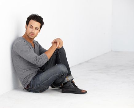 Cool and casual. A handsome young man sitting against a wall while looking away.