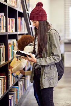 Open a book and you open your mind. a young woman studying from a book while standing by a library bookshelf.