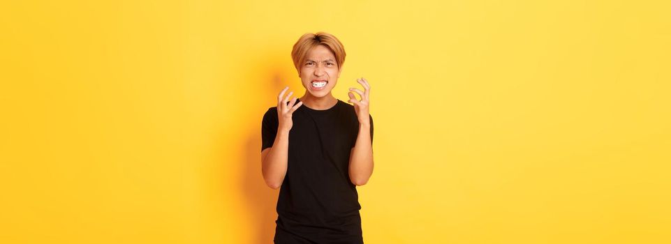 Portrait of angry and pissed-off asian blond guy, clenching hands furious and grimacing, standing over yellow background