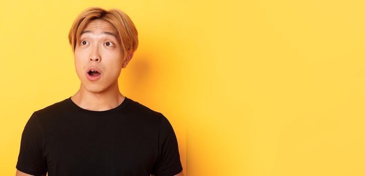 Close-up of impressed asian handsome guy, looking upper left corner with dropped jaw and amazement, standing over yellow background
