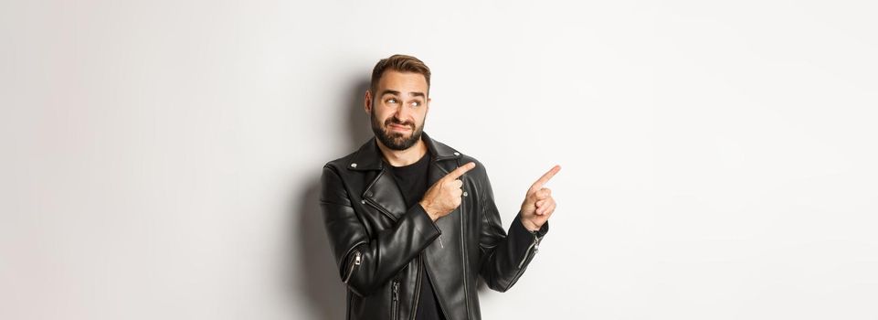 Skeptical and doubtful guy in black leather jacket, shrugging while pointing at upper left corner promo offer, standing over white background