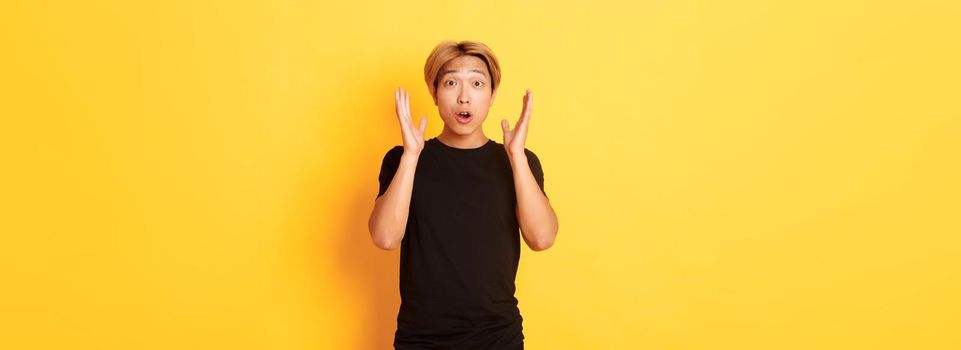 Portrait of surprised gasping blond asian guy reacting to awesome news, yellow background