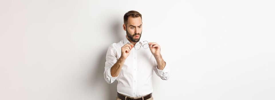 Man looking confused at his glasses, standing in office clothing against white background
