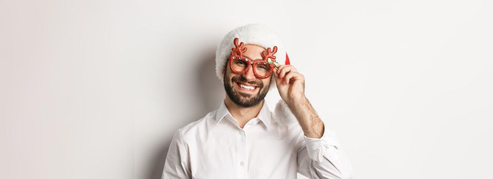 Close-up of handsome bearded guy in xmas party glasses and santa hat, smiling and wishing merry christmas, standing over white background