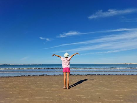 Girl with hat opening her arms in front of the sea, on a sandy beach.