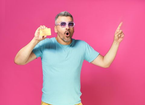 Look at this great offer, grey haired man hold debit, credit card in hand pointing sideways wearing blue t-shirt and purple glasses isolated on pink background. Financial, banking concept