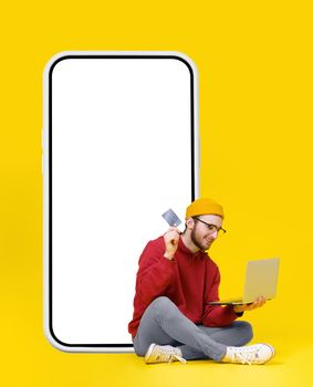 Sitting next to the big smartphone with white screen young man hold laptop and debit or credit bank card in hands wearing red hoodie and yellow hat buying or paying online. Freelancer young man