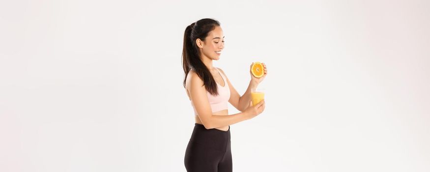 Sport, wellbeing and active lifestyle concept. Portrait of smiling healthy and slim asian girl advice eating healthy food for breakfast, gain energy for good workout, squeezing orange juice in glass
