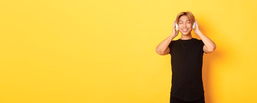 Relaxed and pleased handsome asian guy close eyes from satisfaction while listening music in wireless headphones, standing yellow background