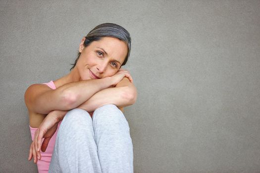 Calm and stress-free. Portrait of an attractive mature woman in gymwear sitting against a gray wall.
