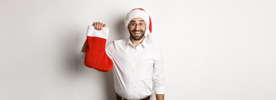 Merry christmas, holidays concept. Excited bearded guy in santa hat holding xmas sock and smiling, celebrating New Year, white background