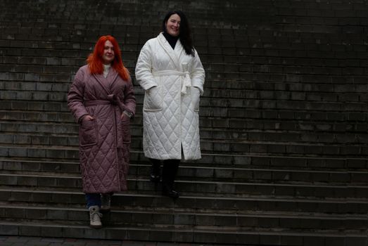 Two plus-size women posing in coats against a staircase backdrop.
