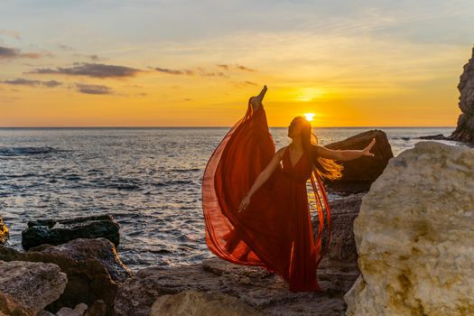 a dancing woman in a red flying dress on the ocean or on the sea beach against the backdrop of the sunset sky.
