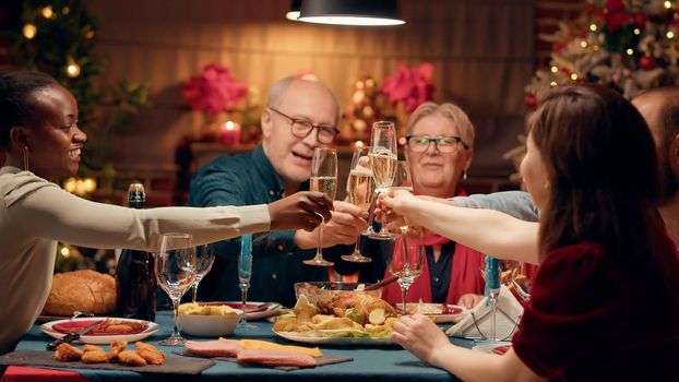 Positive festive people celebrating Christmas at evening dinner while clinking glasses with champagne
