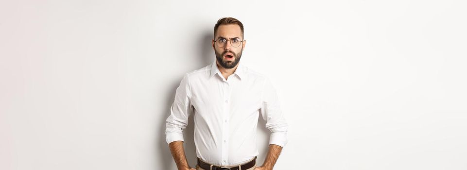 Shocked and displeased businessman in glasses, gasping and looking upset at camera, standing over white background
