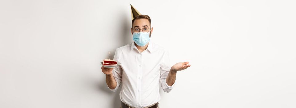 Coronavirus, quarantine and holidays. Confused man in face mask, holding birthday cake and shrugging, standing over white background clueless
