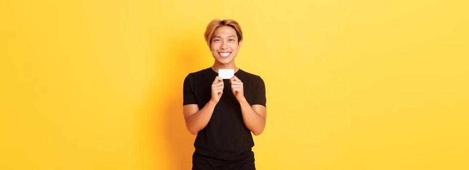 Excited happy asian guy smiling and showing credit card, standing yellow background