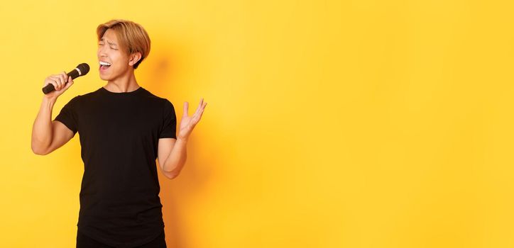 Carefree handsome asian guy, korean singer singing into microphone with passion, standing yellow background
