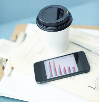 Coffee helps the brain make sense of the numbers. Smartphone with a bar chart on the screen sitting ontop of a newspaper and clipboard, with a disposable cup and pen.