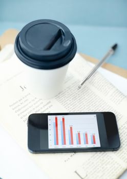 Portable office. Smartphone with bar char on screen sitting ontop of a newspaper and clipboard with a disposable cup and pen.