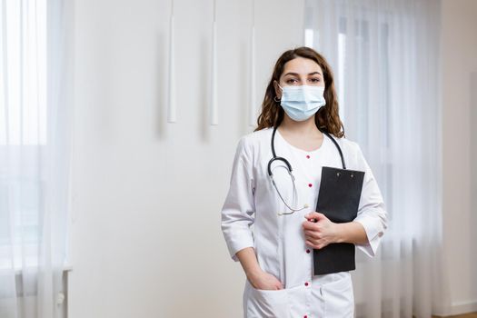 Portrait of young female doctor in medical mask in white coat with stethoscope, holding folder with documents, looking at camera, quarantine