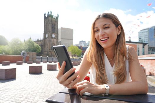 Smiling girl with mobile phone in her hands sits at the table outdoors with the cityscape on the background. Beautiful young woman is watching videos and photos on her smartphone at the cafe table.