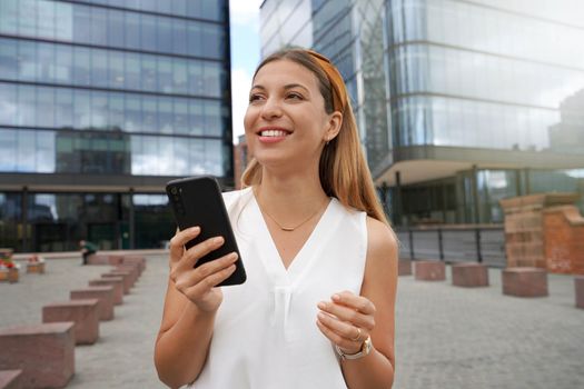 Portrait of a beautiful young female entrepreneur looking away holding a smartphone against business buildings. Charming young woman going to work.
