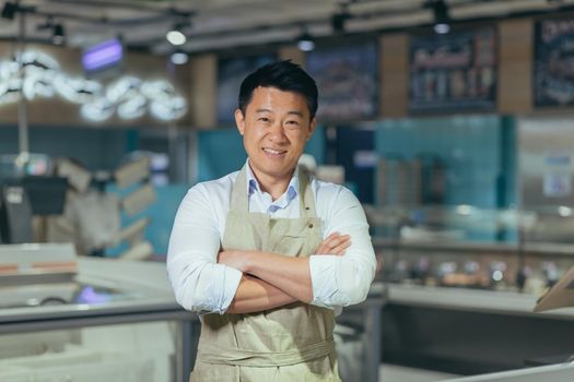 portrait of handsome asian man salesman in apron standing and looking at camera in grocery store supermarket. Male worker seller or small business owner. indoor. food, groceries market Cheerful smile.