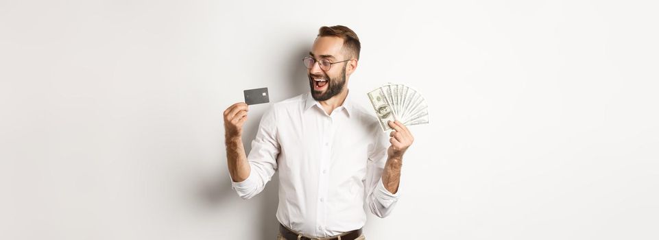 Excited businessman holding money and looking at credit card, standing over white background