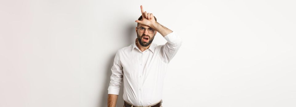 Shocked guy showing loser sign on forehead, complaining, standing over white background