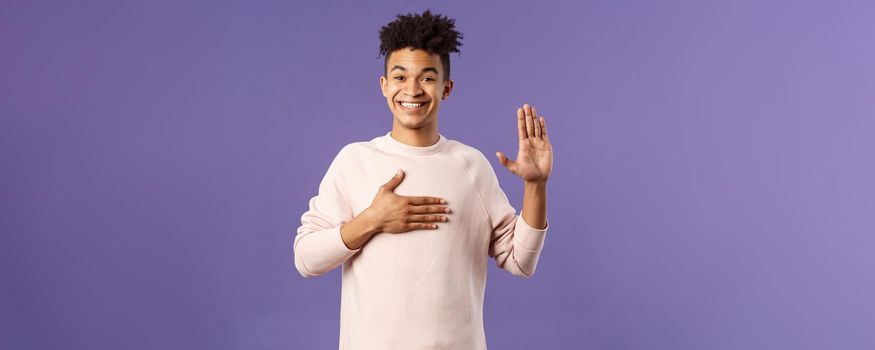 Portrait of young happy, joyful hispanic man making honest statement, promise to tell only truth, hold one hand on heart and another raised, smiling sincere, standing purple background