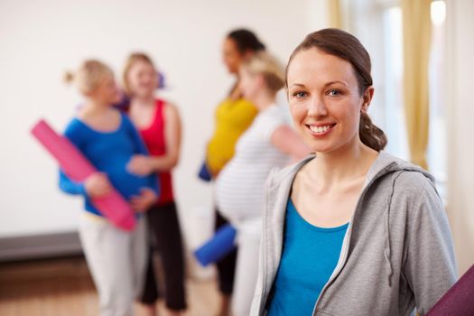 Relaxation exercise in perfect for pregnancy. A young pregnant woman standing in a gym holding an exercise mat with a group of women in the background.