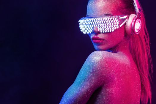Beautyful girl with glitter and sparkles on her face and body. Portrait of sexy TDJ with headphones and neon sunglasses