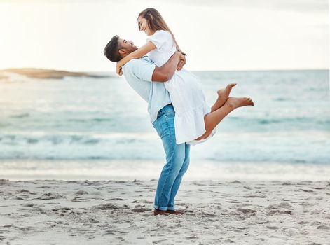 Man at beach lift woman with love, smile with sun setting over the horizon or background. Young couple travel to ocean on vacation, happy together with sunset over sea or waves in nature backdrop.