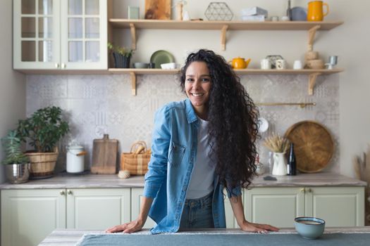 Portrait of happy hispanic woman at home in kitchen, woman with curly hair smiling