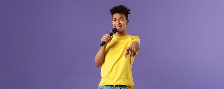 This song is for you. Portrait of romantic carefree hispanic man singing karaoke, pointing at camera as dedicate his performance, holding microphone, standing purple background