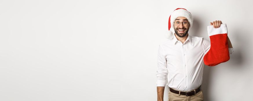 Merry christmas, holidays concept. Excited bearded guy in santa hat holding xmas sock and smiling, celebrating New Year, white background