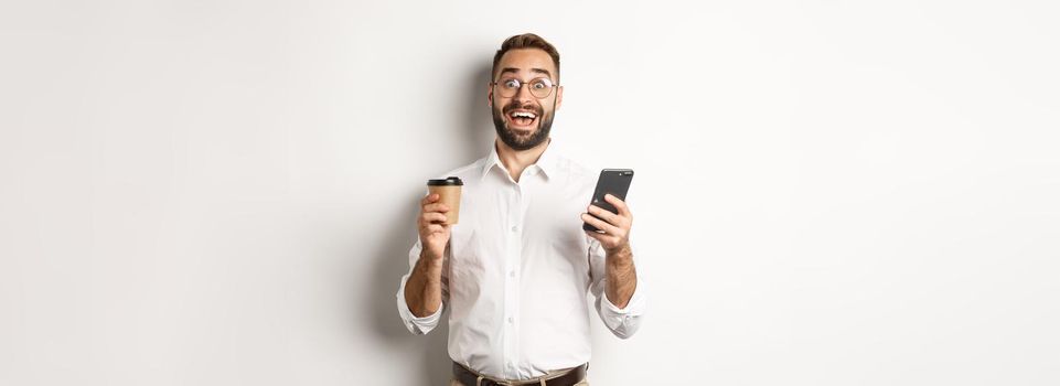 Amazed businessman drinking coffee, reacting at awesome online offer on mobile phone, standing over white background