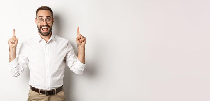 Happy male entrepreneur in business clothes, pointing fingers up and showing promo offer, standing over white background