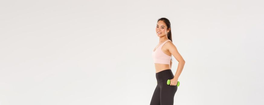 Sport, gym and healthy body concept. Full length of sweaty smiling female athlete, cute slim girl asian workout with dumbbells, lifting weight over white background, training in gym, white background
