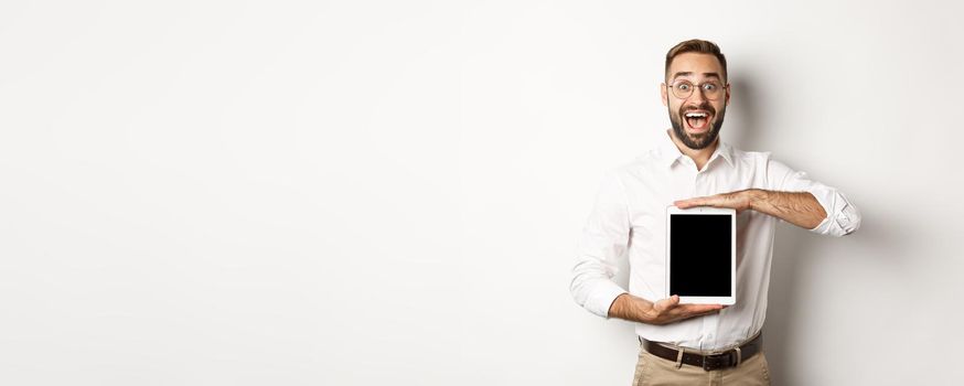 Excited man showing digital tablet screen, smiling amazed, standing over white background