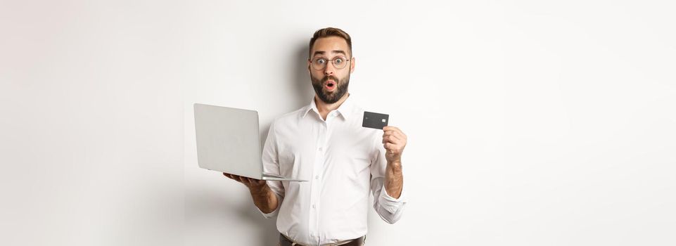 Online shopping. Surprised man holding laptop and credit card, shop internet store, standing over white background