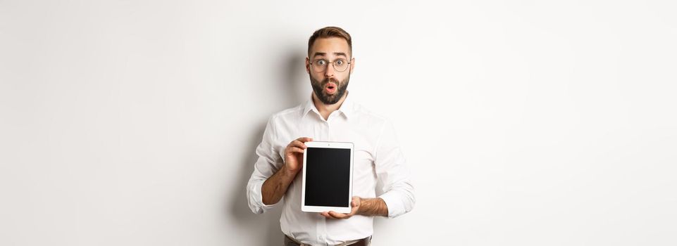 Surprised man in glasses, showing digital tablet screen, looking amazed, standing over white background