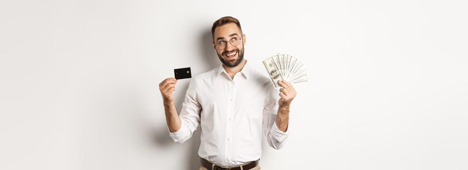 Image of young businessman holding credit card and money, looking at upper left corner and thinking about shopping, standing over white background