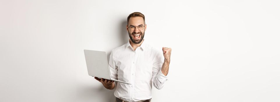 Business. Happy manager winning online, rejoicing with fist pump, holding laptop and triumphing, standing over white background