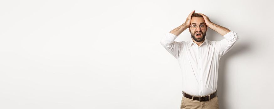 Frustrated businessman holding hands on head, looking shocked and anxious, standing over white background