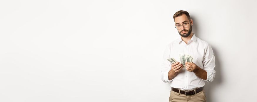 Handsome businessman counting money and looking at camera, standing serious against white background