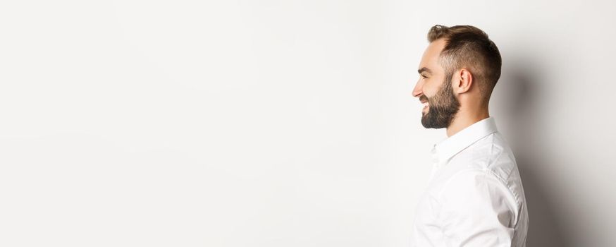 Close-up profile shot of handsome bearded man looking left and smiling, standing against white background