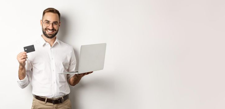 Online shopping. Handsome man showing credit card and using laptop to order in internet, standing over white background