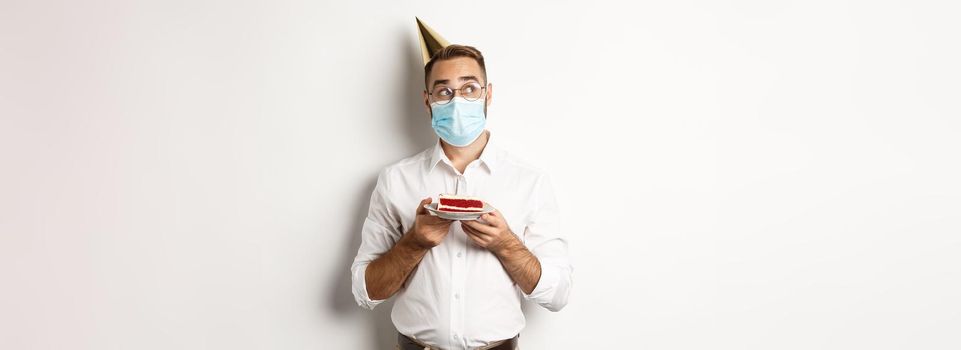 Covid-19, social distancing and celebration. Thoughtful man holding birthday cake, making wish and wearing face mask on quarantine, white background